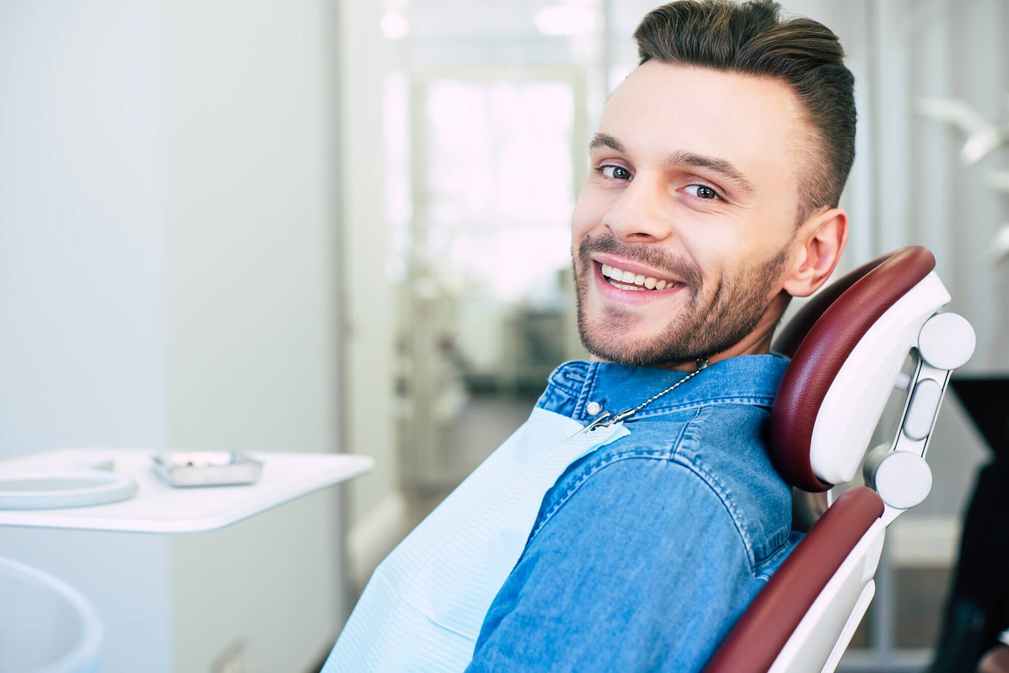 Happy man with hazel eyes and dark brown hair is sitting in dental chair and smiling right into the camera because he is satisfied with the work of a dentist